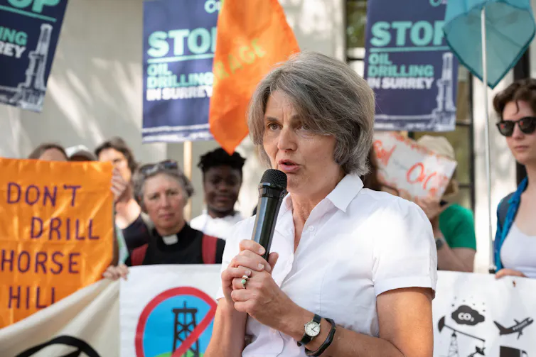 woman in white shirt, grey hair, microphone with colourful stop fossil fuel banners being held in background