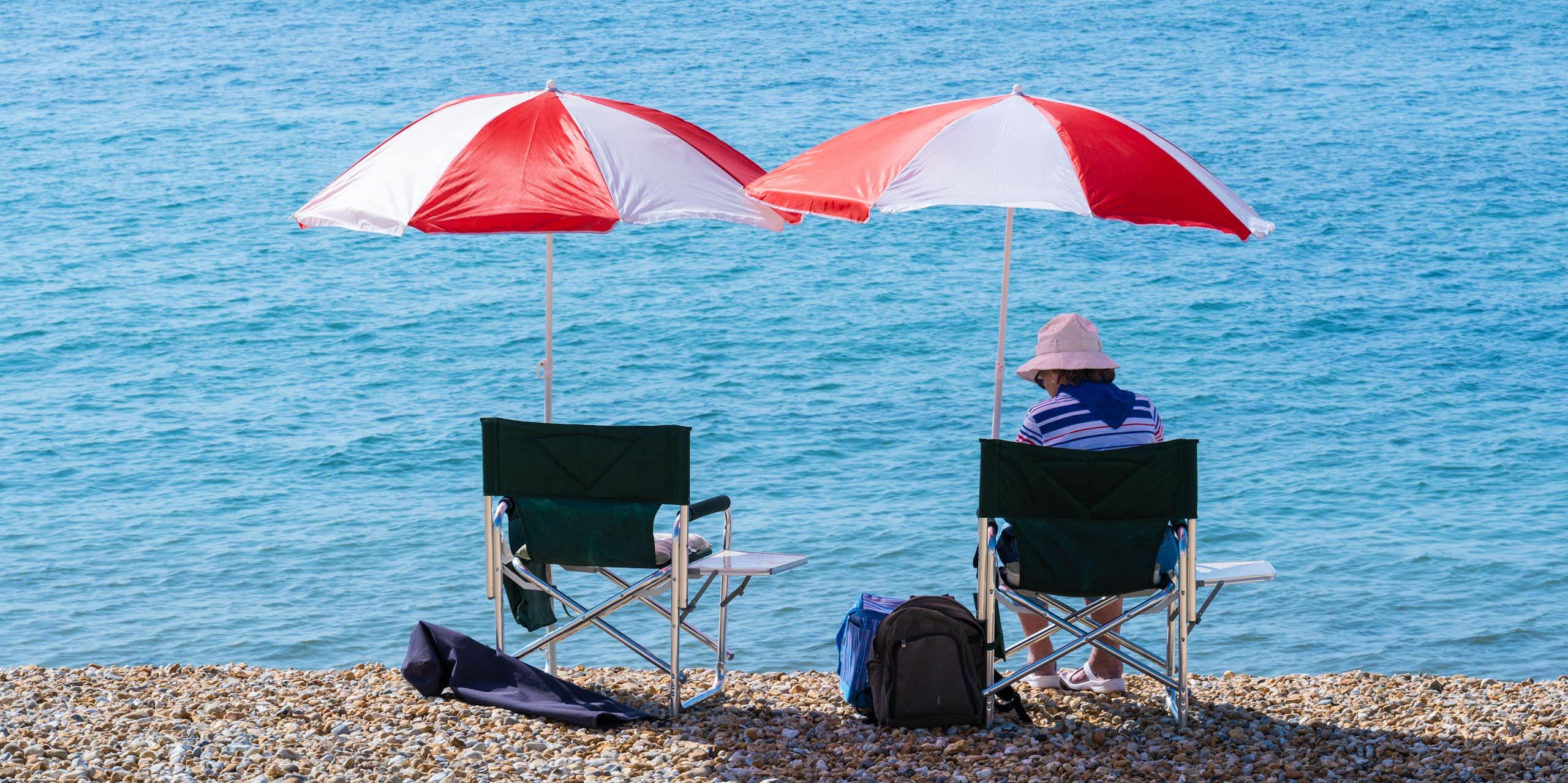 A person sitting on a pebbled beach with a red and white umbrella overhead