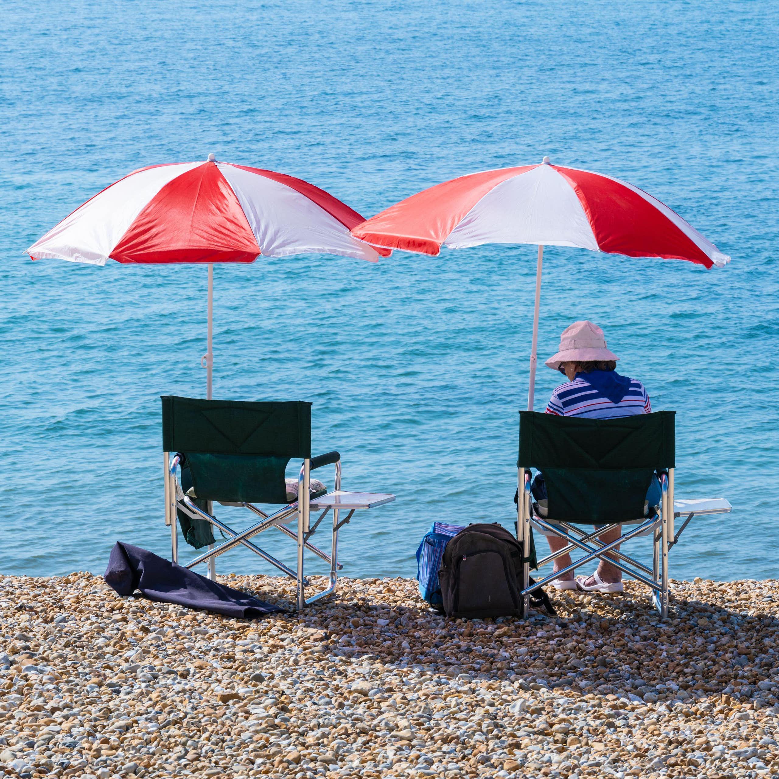 A person sitting on a pebbled beach with a red and white umbrella overhead