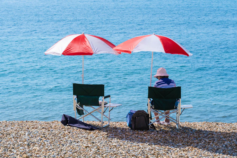 A person sitting on a pebbled beach with a red and white umbrella overhead