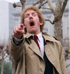 A man dressed in a mac with a horrified and distressed look on his face.