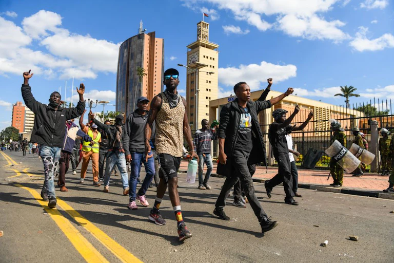 Young men walk on a road, some holding up arms or raising fists, as police in riot gear stand on the side of the road