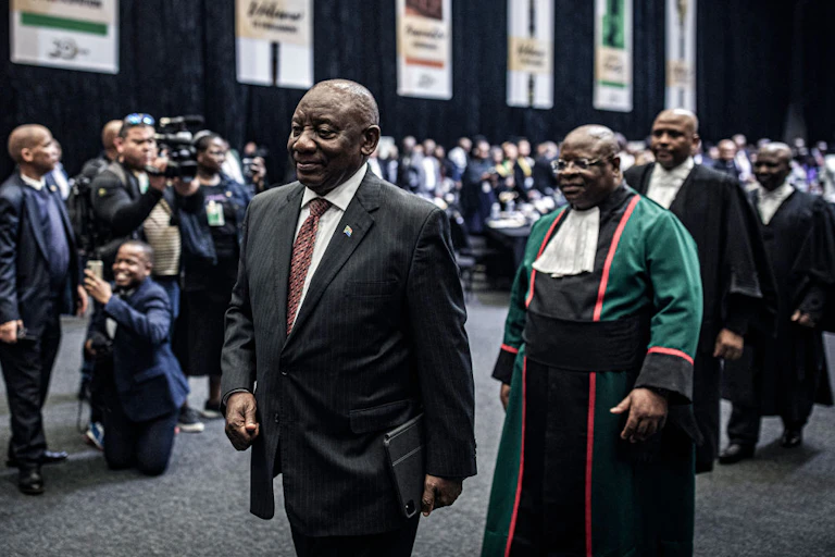 Man in suit and tie walks ahead of a procession of men in gowns, with photographers nearby and more people in the background