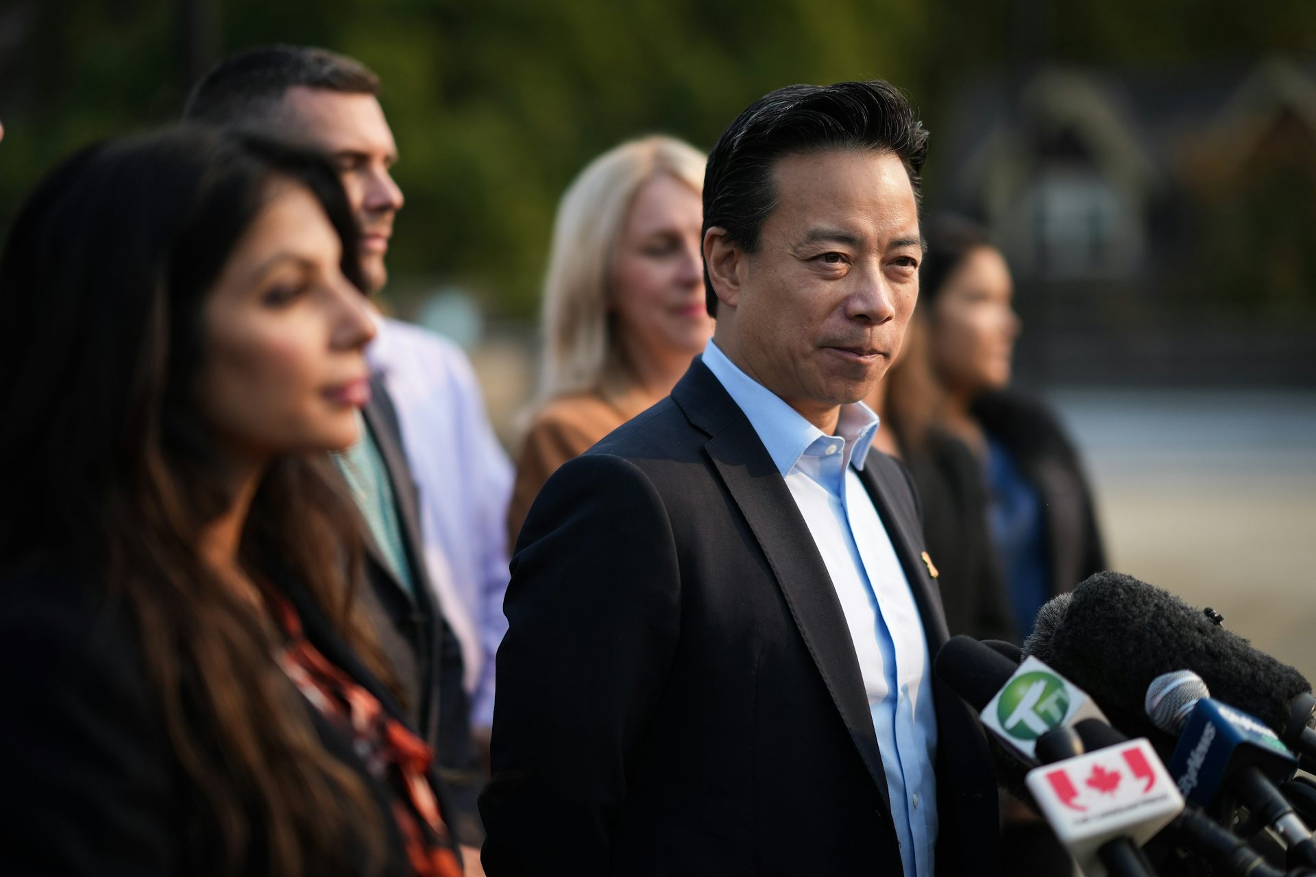 An Asian man stands behind a bank of microphones with other people standing around him.