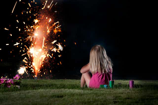 A young girl is seen sitting on a lawn, watching fireworks from a distance.