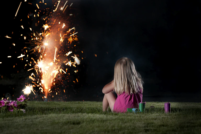 A young girl is seen sitting on a lawn, watching fireworks from a distance.
