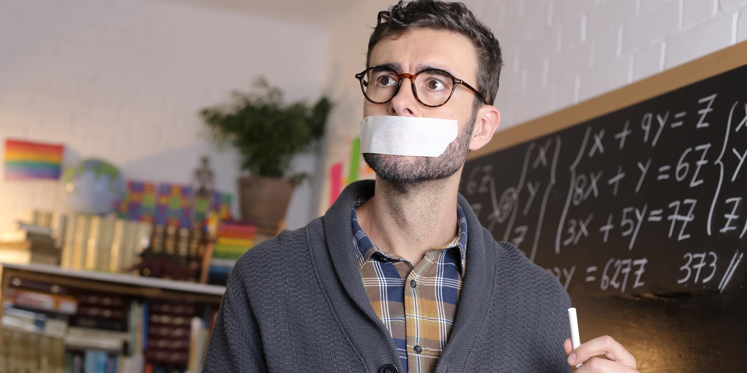 A professor wearing a plaid shirt and sweater holds a piece of chalk while a piece of tape covers his mouth. Math equations are on a blackboard in the background. 