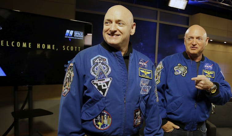 Two identical men wearing blue jumpsuits stand next to each other.