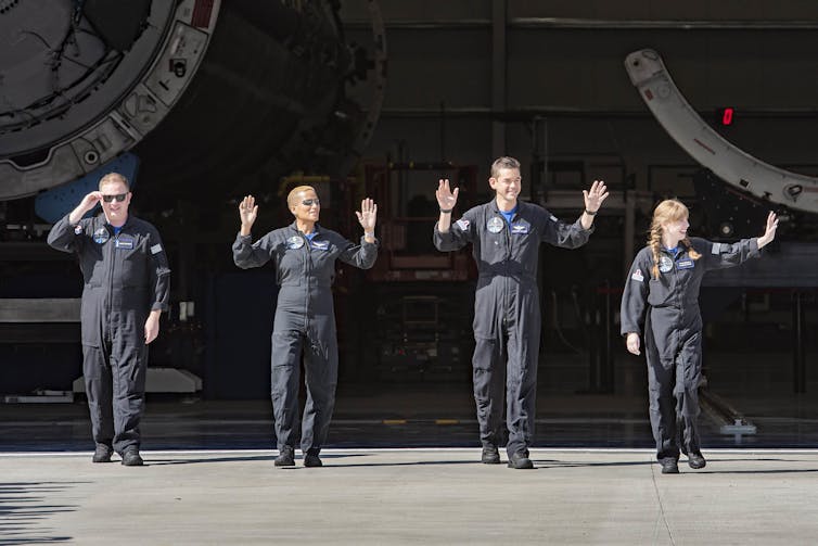 Four people wearing black jumpsuits wave their hands in the air.