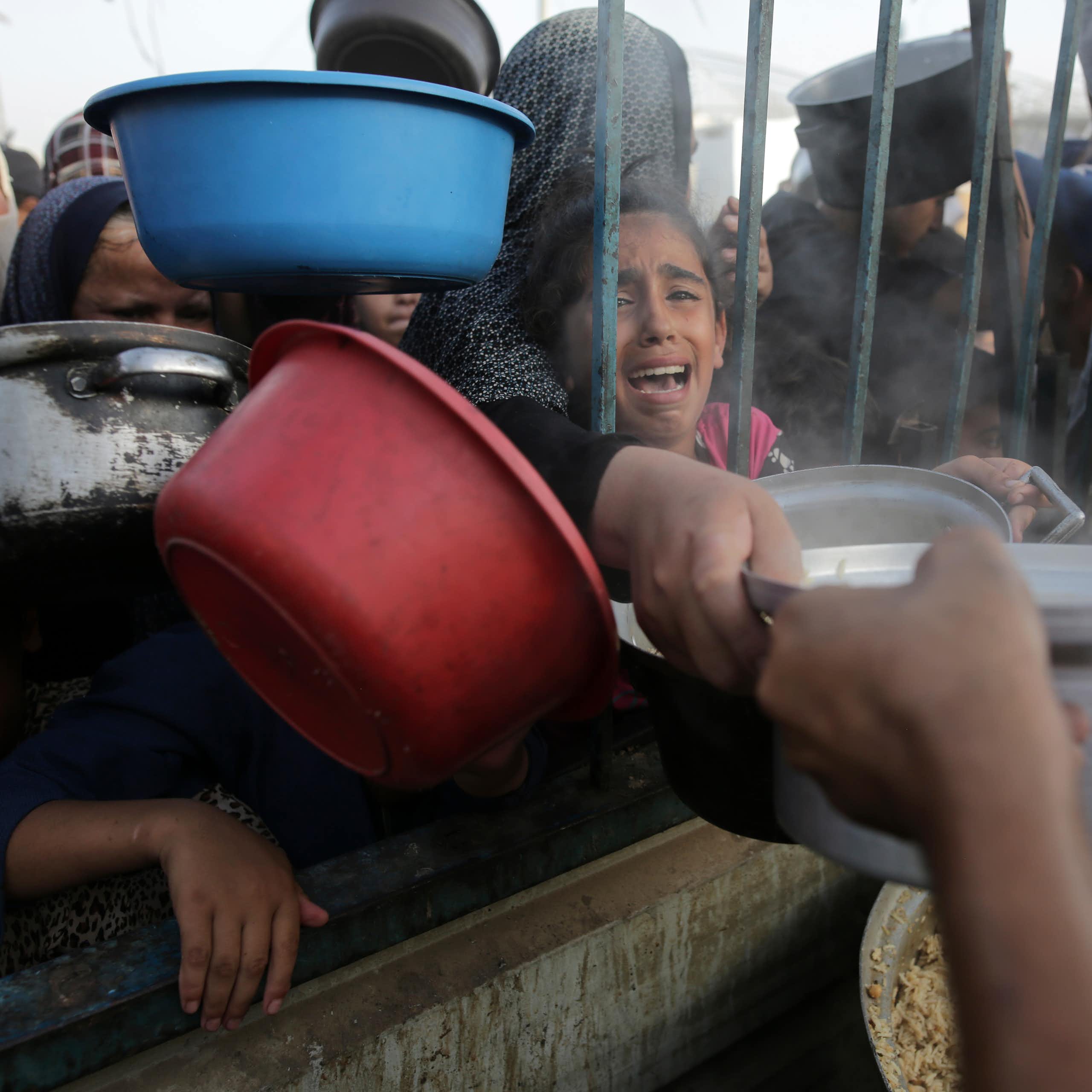 A young girl cries as she reaches through bars for food, as other people clamor around her with bowls.
