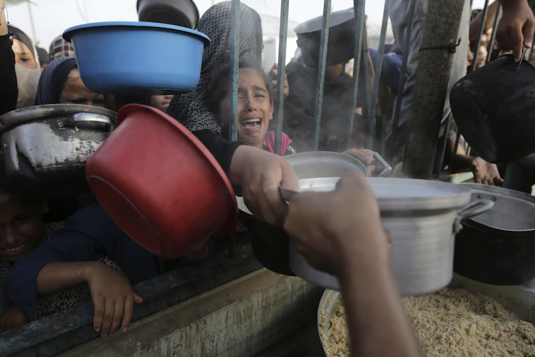 A young girl cries as she reaches through bars for food, as other people clamor around her with bowls.
