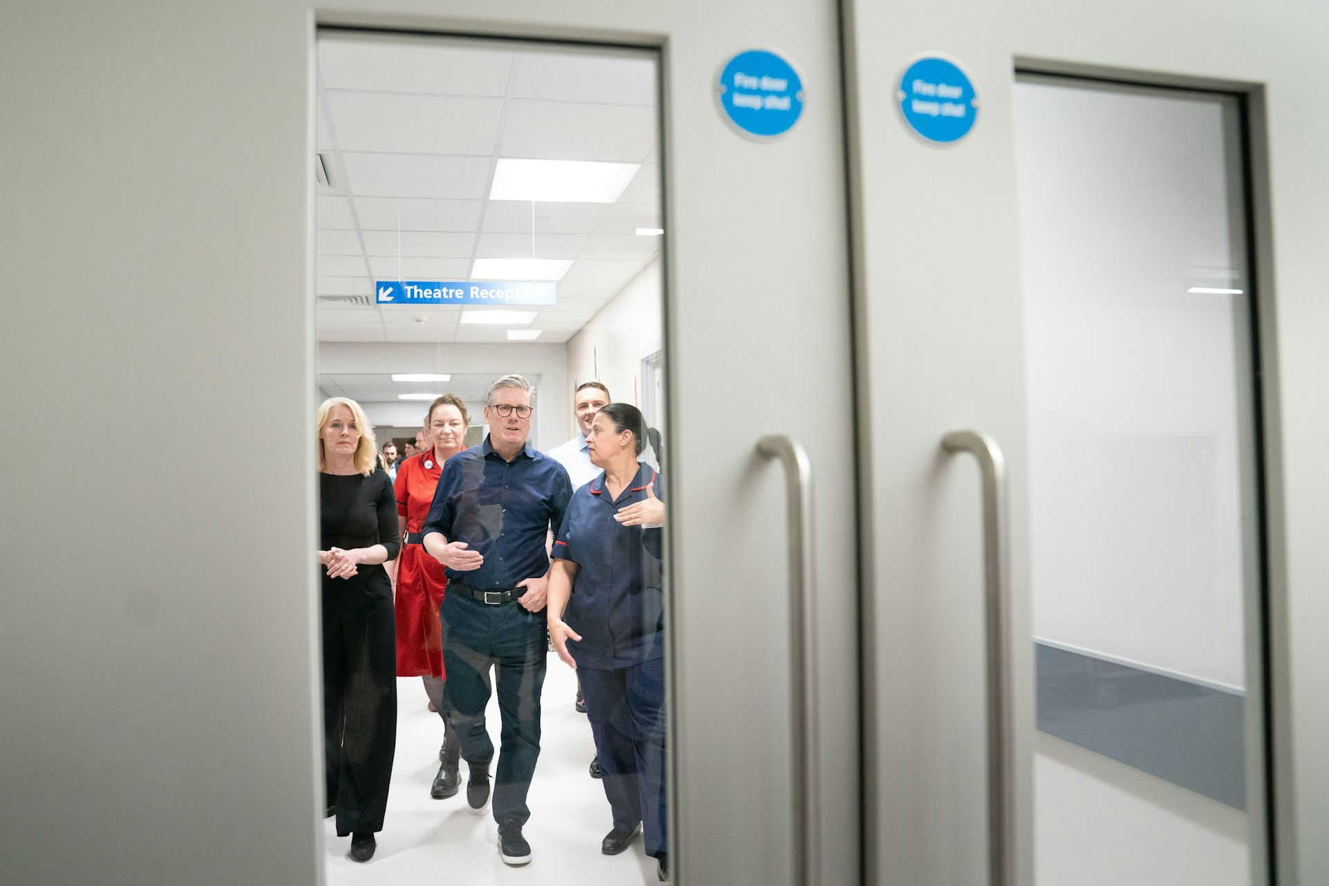 Vista através de um painel de janela de corredor de Keir Starmer caminhando com funcionários de um hospital