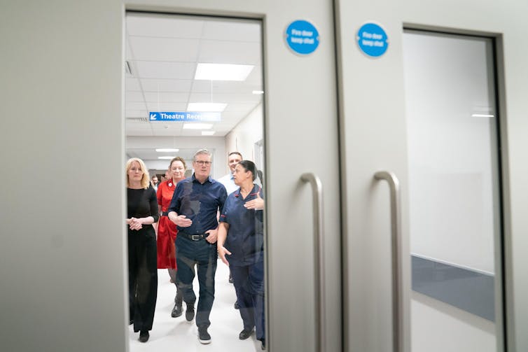 Vista através de um painel de janela de corredor de Keir Starmer caminhando com funcionários de um hospital