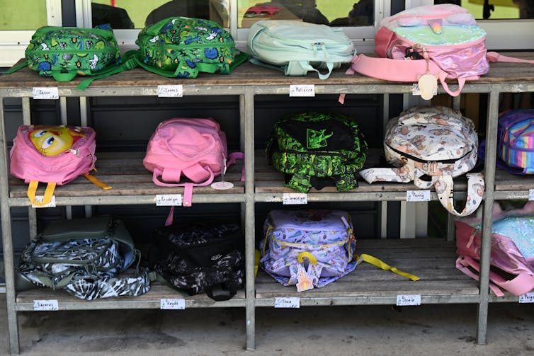 Backpacks lie on shelves, next to name tags, outside a school classroom.