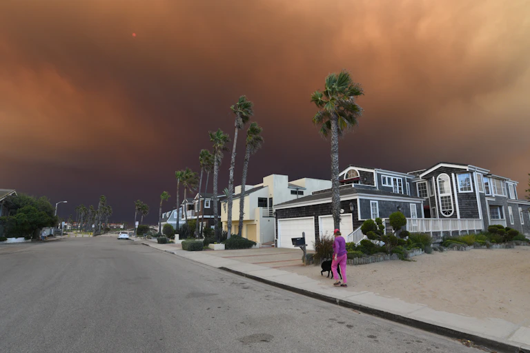 A person walks a dog in a residential neighborhood of Oxnard as orange wildfire turns the sky dark overhead. The street is lined with palm trees and is otherwise empty...
