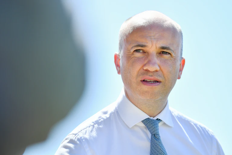Matt Kean speaks to the media in Sydney, against a pale blue sky, with a grey shadowed figure on the left of the frame