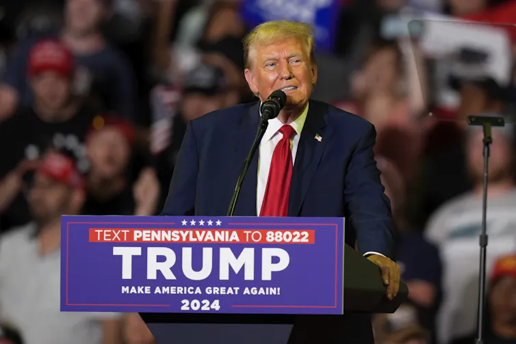 A man in a suit stands at a lectern that reads TRUMP 2024