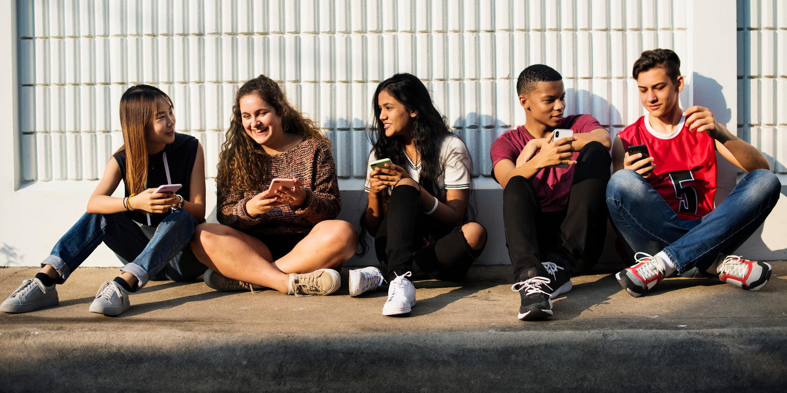 Five diverse teens sitting on the ground socialising over smartphones.