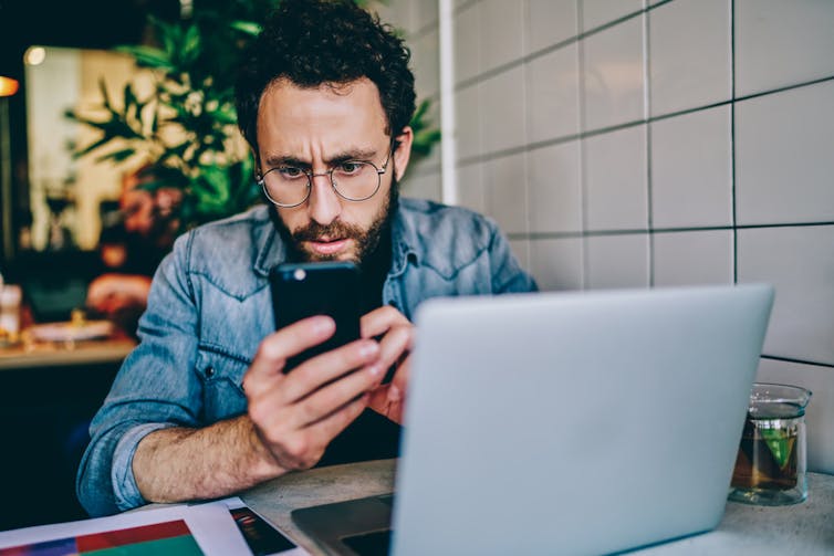A man with a black beard and glasses looks at his smartphone while frowning, sitting at a cafe with his laptop.