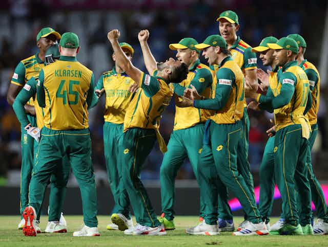 A sports team in green and gold, players grouped together smiling and clapping, a man in the middle raises his fists in celebration.