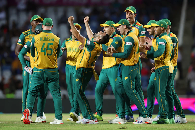 A sports team in green and gold, players grouped together smiling and clapping, a man in the middle raises his fists in celebration.
