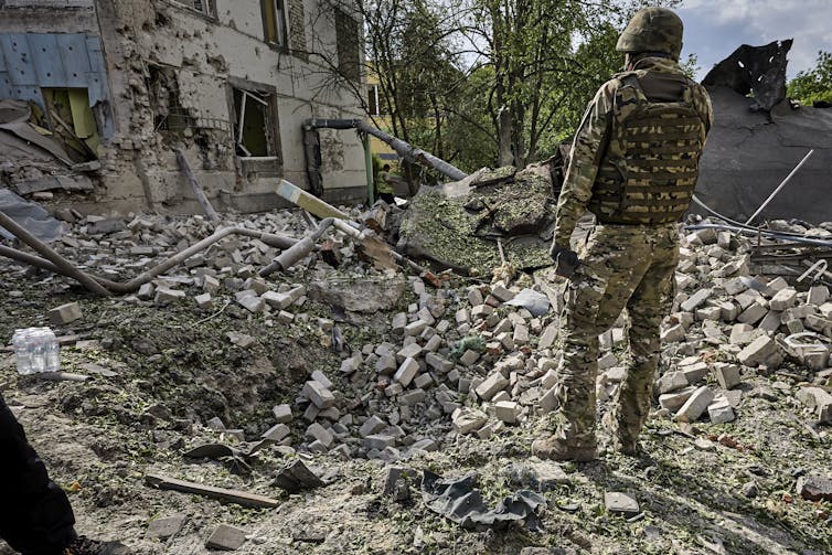 A man in camouflage stands surrounded by rubble at the site of a bomb attack.