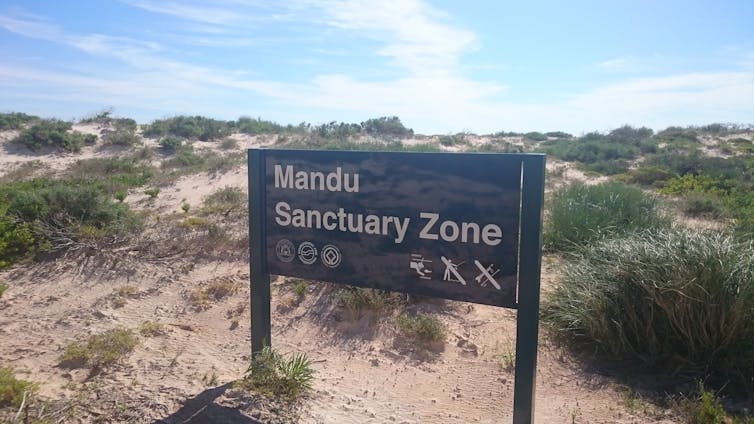 brown sign stating Mandu Sanctuary Zone, with grassy sand dunes in background, blue sky