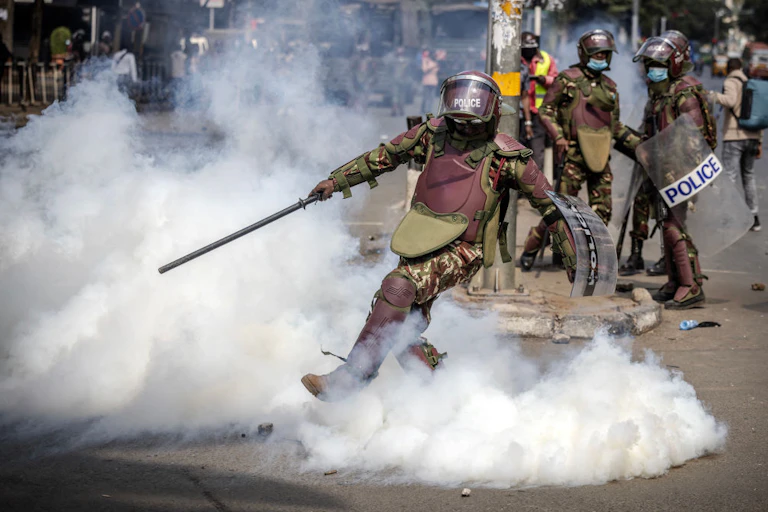 A man dressed in brown and green fatigues and wearing a helmet kicking his foot into a cloud of smoke.