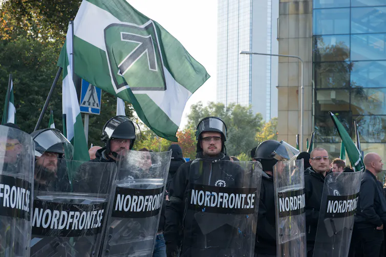 A line of black-clad men stand on a street, wearing helmets and holding police-style shields.