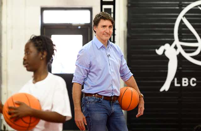A dark-haired man holds a basketball with a Black youth in front of him looking away and also holding a basketball.