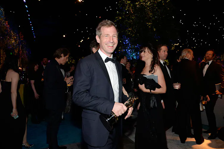 Young man wearing tuxedo smiles as he holds a gold statuette.