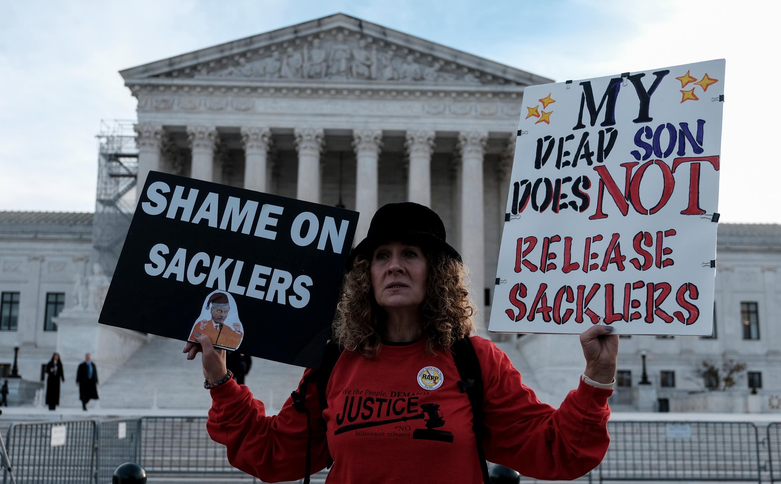 Woman in red sweatshirt holds signs saying 'Shame on Sacklers' and 'My dead son does NOT release Sacklers."