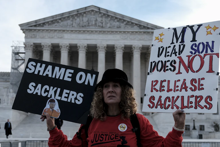 Woman in red sweatshirt holds signs saying 'Shame on Sacklers' and 'My dead son does NOT release Sacklers."