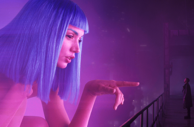 A massive holograph of a pink-skinned, blue-haired young woman points at a small man who gazes up at her.