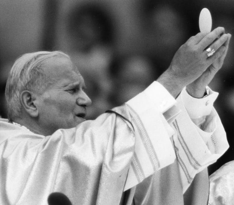 An elderly man in white clothes and a cap raises his hands as if praying.