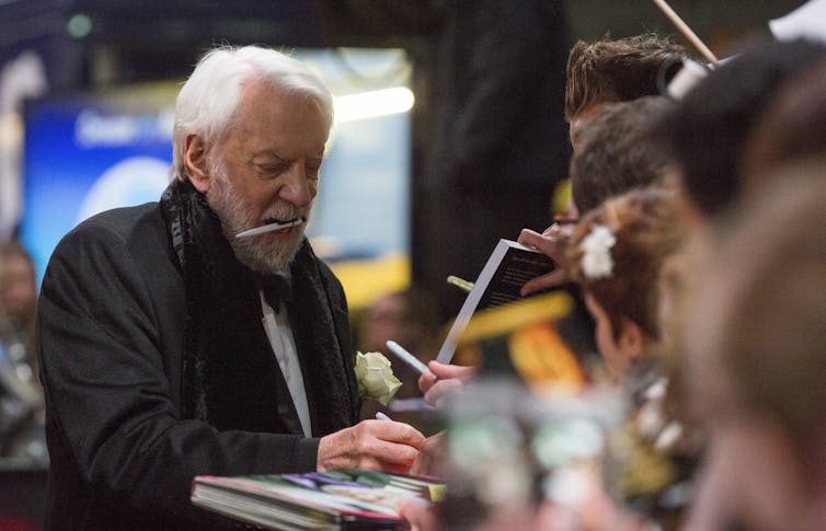 A man with a pen in his mouth signs autographs as fans lean in.