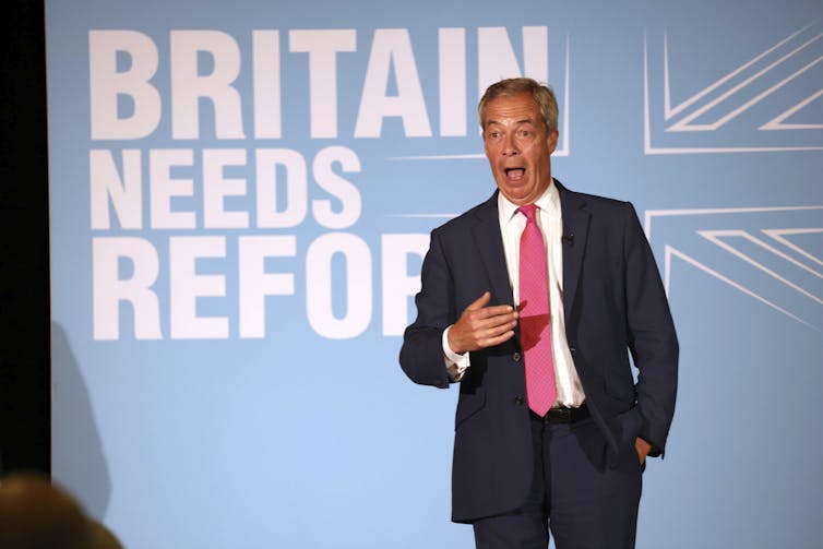 Nigel Farage stands in front of a banner calling for reform.