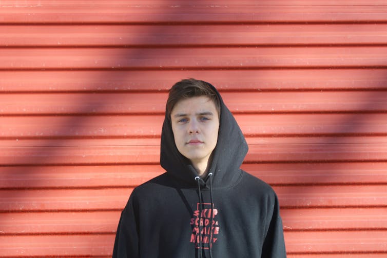 A young man stands against a red timber wall. He wears a black hoodie jumper.