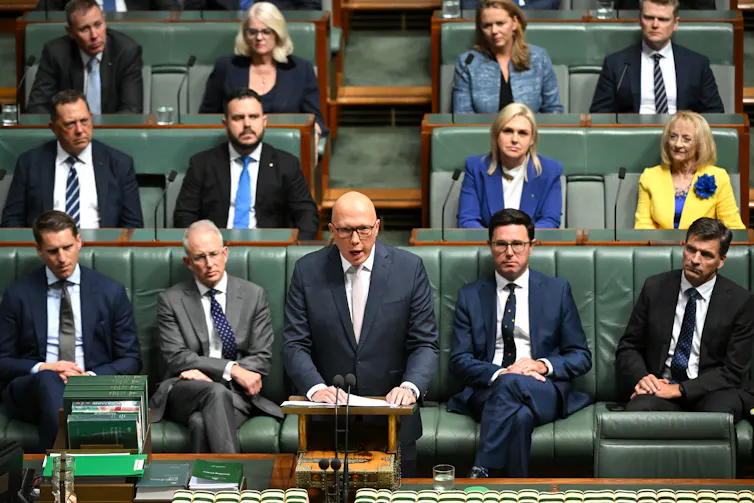 A man in a suit stands at a lectern in front of many others sitting in tiered seating.
