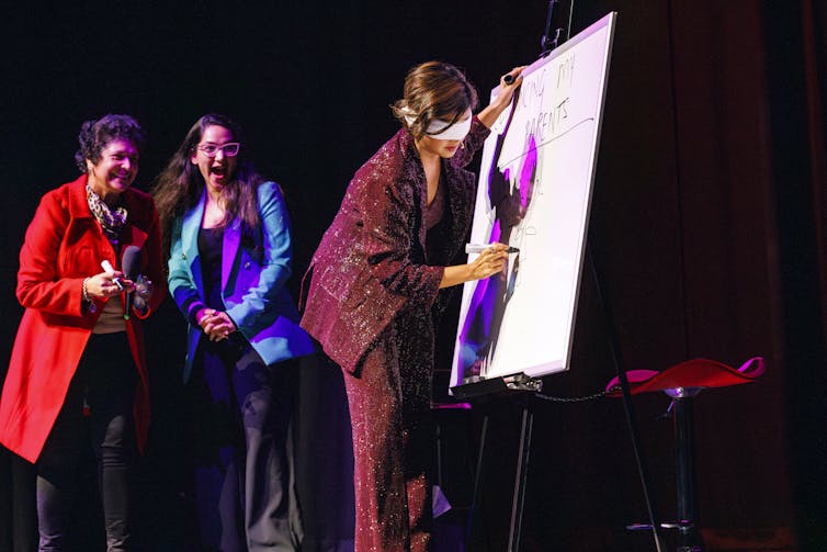 Two women on stage, watching a blindfolded Shah draw.
