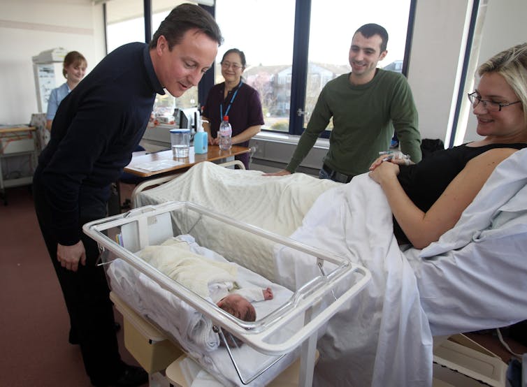 David Cameron looking at a newborn baby in a hospital crib, next to a woman in a hospital bed.