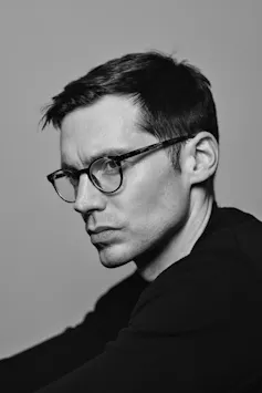 A serious-looking young man dressed in a black sweater with black glasses.