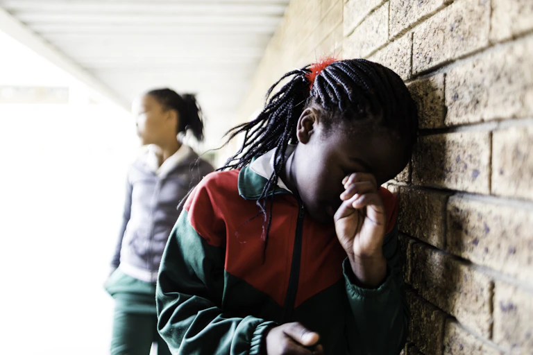 A girl turns her head towards a wall, holding a hand to her face. Another girl stands in the background.