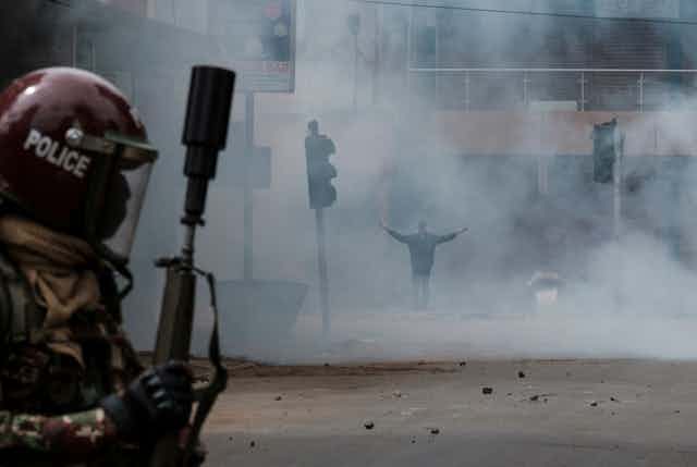 A man in the background standing on a street enveloped in a white cloud with his hands raised, while a man in the foreground holds a tear gas launcher