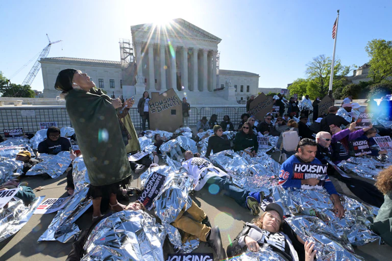 Around one or two dozen people lie on the ground in emergency blankets, some holding signs, outside a building with tall white pillars.