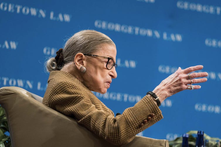 A woman wearing eyeglasses is sitting in a chair and raises her hand to make a point.