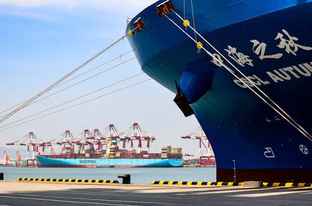 Two container ships -- one, dark blue, in the foreground, and another, light blue, in the distance -- are seen amid port infrastructure.