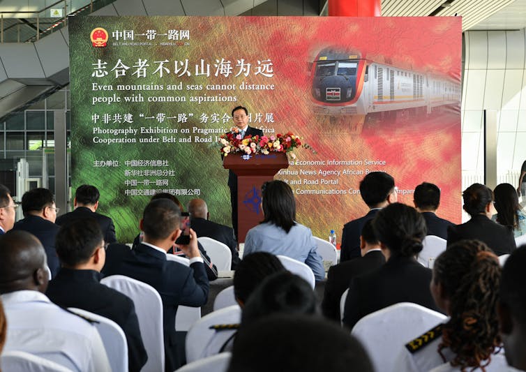 Xu Jianping, a Chinese official, stands behind a podium, speaking to a room full of professionals in business dress. Behind him, a sign in Chinese and English reads, in part,