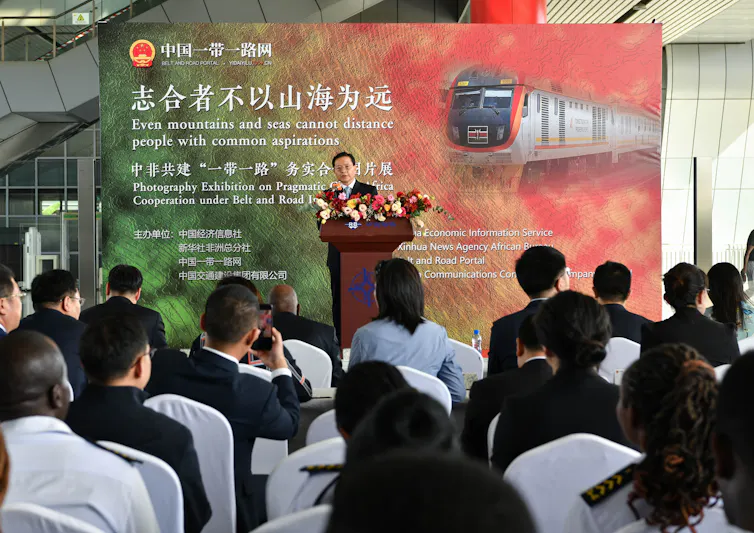 Xu Jianping, a Chinese official, stands behind a podium, speaking to a room full of professionals in business dress. Behind him, a sign in Chinese and English reads, in part,