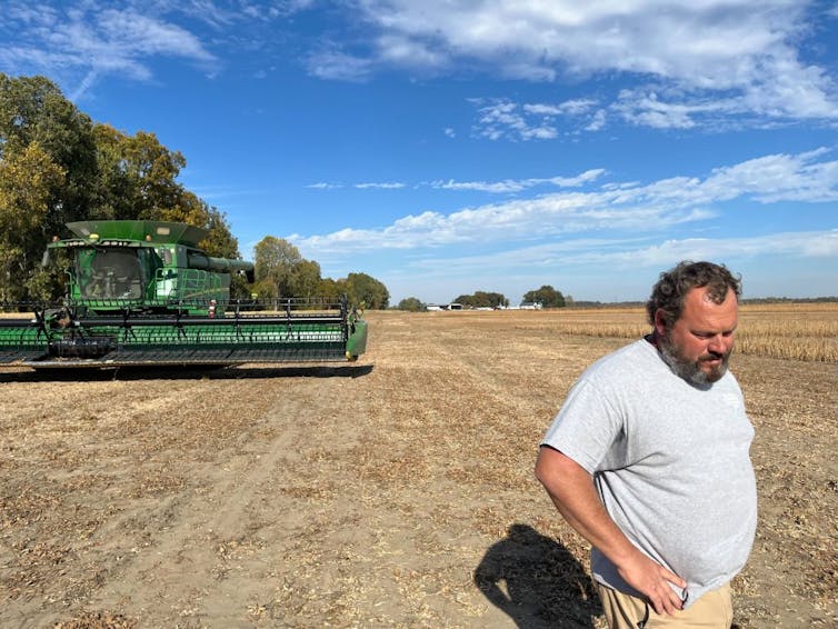 Man with beard takes a break from harvesting soybeans on a farm in Tennessee. A green soybean harvester idles in the background.